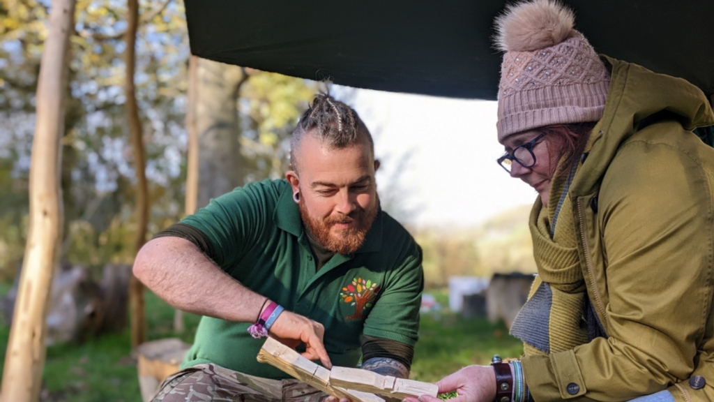 Image of two people sitting under a canopy in a wooded area. They are carving small pieces of wood. Credit: Raincliffe Wood Community Enterprise CIC