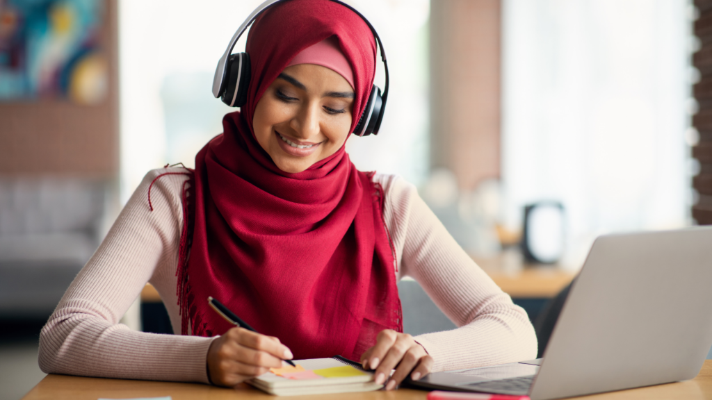 Woman of colour wearing a Hijab head scarf sat with headphones on in front of an open laptop. She is writing in a notebook. 