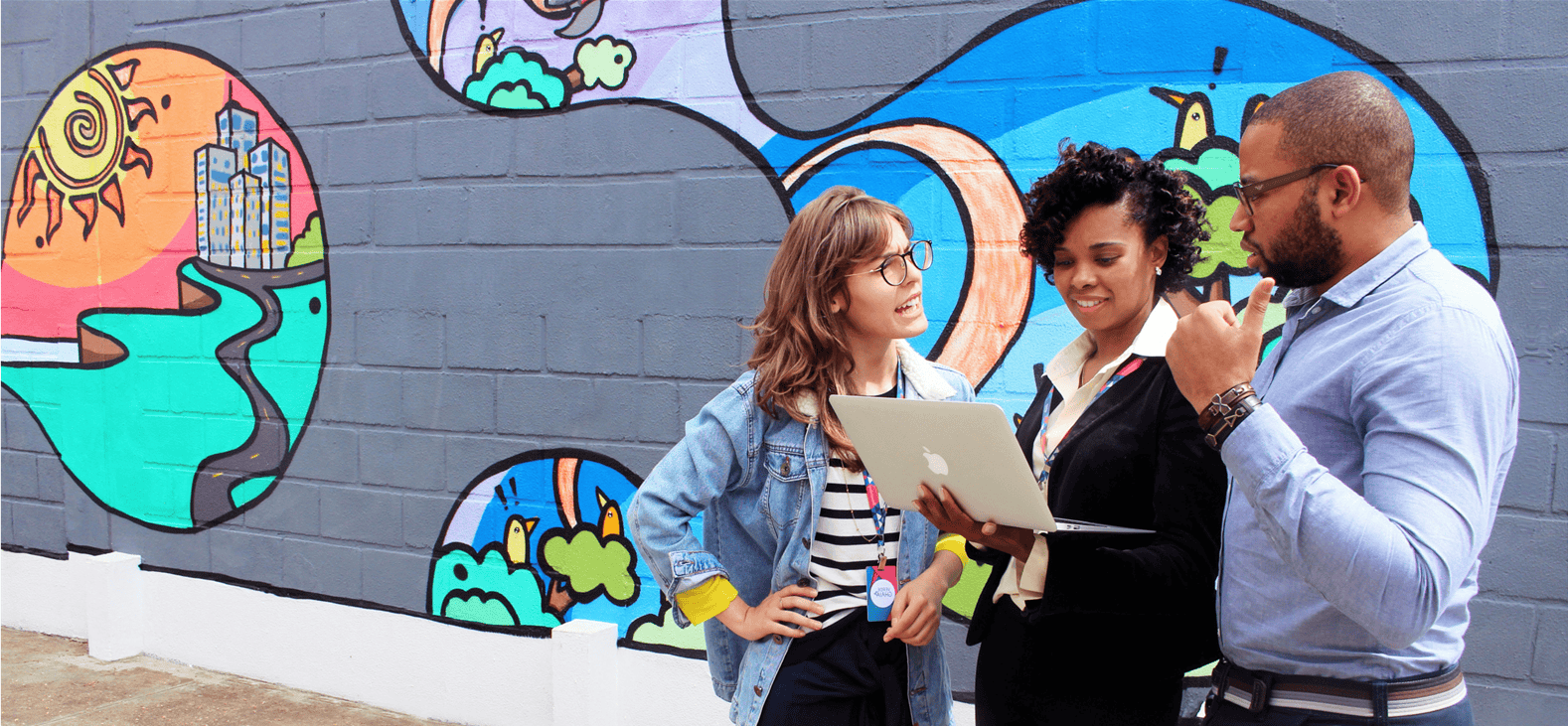 A group of three people stand in front of a wall covered in brightly coloured graffiti all looking at one laptop and discussing what's on the screen.