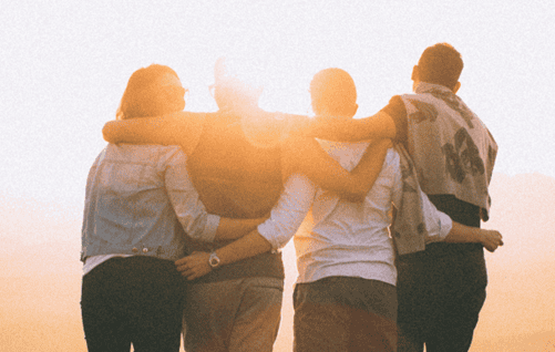 Four people with their backs to the camera watching a sunset with their arms around each other.