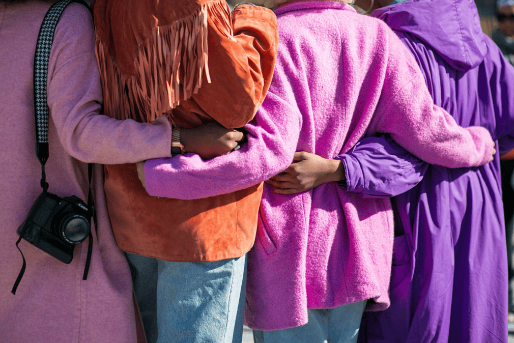 The back of four women wearing different coloured coats and linking arms around each other. The coats are pinks, purples and orange and one is carrying a camera on their shoulder.