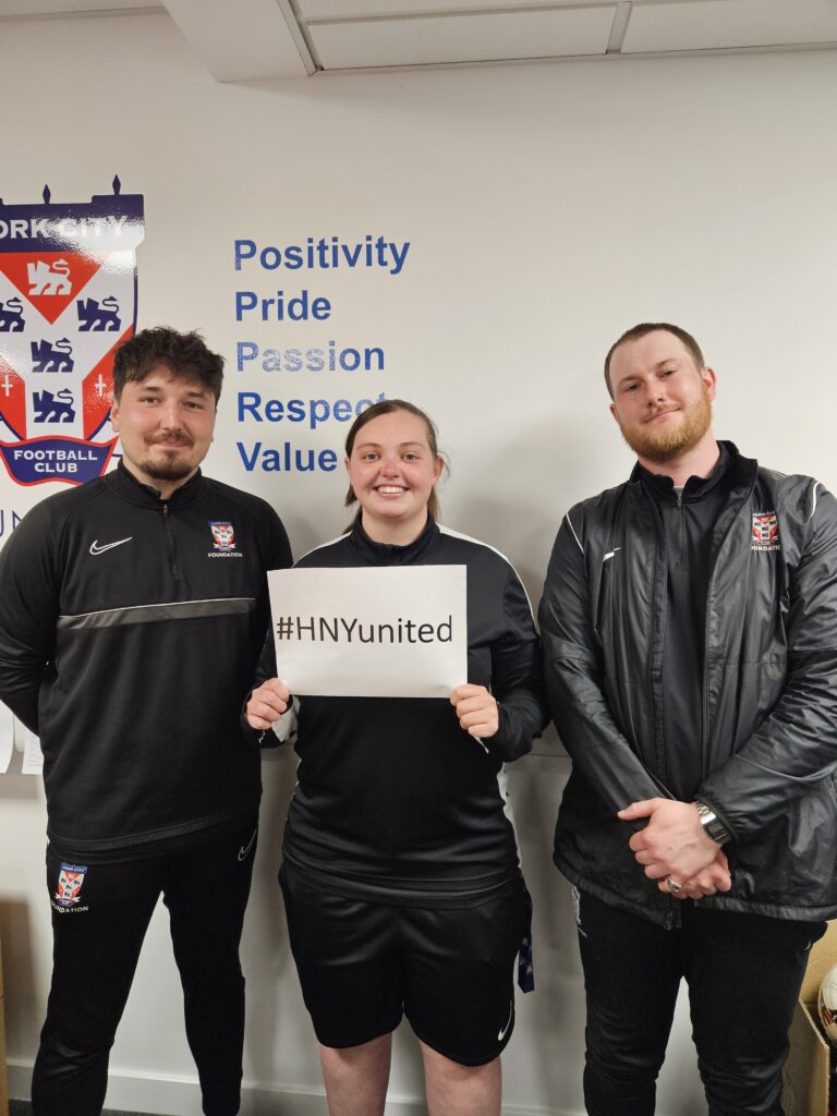 Sam Witthames (left), Lauryn Zareba (middle), Zack Holdaway (right), hold up a sign saying #HNYunited. They are all foundation coaches at York City Football Club Foundation.