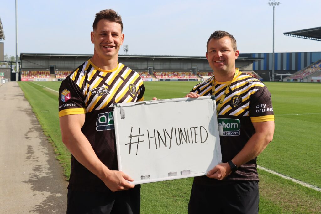 York Knights players Jesse Dee and Bailey Antrobus in their playing kit in the York ground, holding up a whiteboard with the word, #HNY UNITED