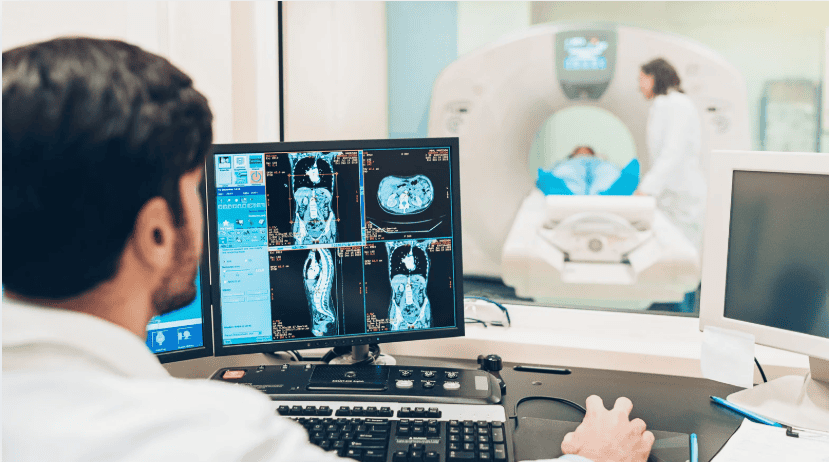 Diagnostic image. A Radiologist sits at the computer with back to camera which in font of them the patient is in the MRI scanner with an assistant by their side.